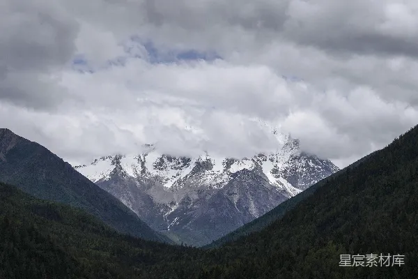 高清摄影 白天阴云密布的天空下，山峦被白雪覆盖，近旁绿树成荫。