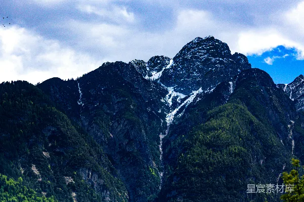 高清摄影 云层密布的天空下，白雪覆盖的山峰旁有绿树环绕。