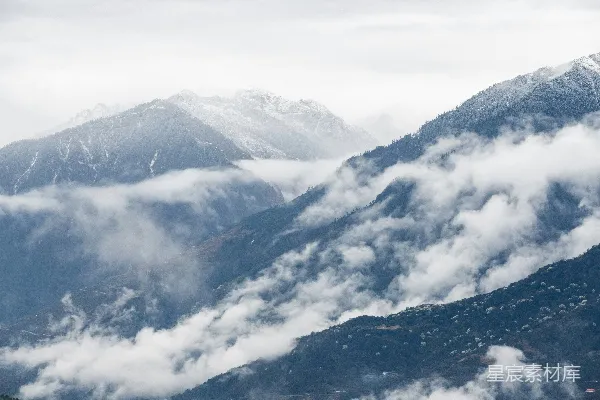 高清摄影 云雾缭绕的山峦下，白雪覆盖的山峰在阴云密布的天空中若隐若现。