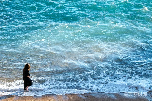 高清摄影 一个女人在阳光下的海滩上漫步，海浪拍打着岸边。