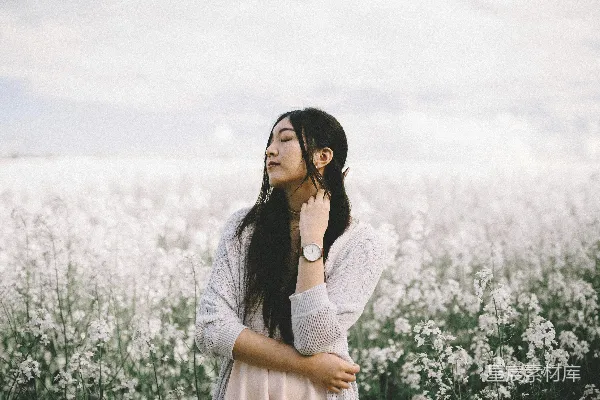 高清摄影 一名穿着桃色内搭和灰色针织开衫的女子站在白天的白色花丛后面拍摄。