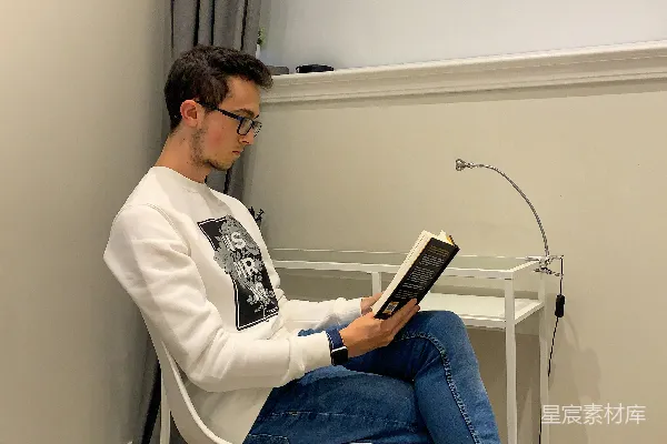 高清摄影 穿白色衬衫和蓝色牛仔裤的男人坐在白色的椅子上阅读一本书。