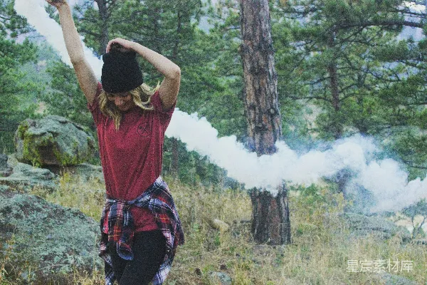 高清摄影 女人双手举高，手持白色烟雾弹，站在岩石旁，周围是树木，在白天。