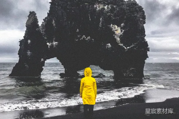 高清摄影 穿黄夹克的男人站在日间海边的岩石形成物附近。